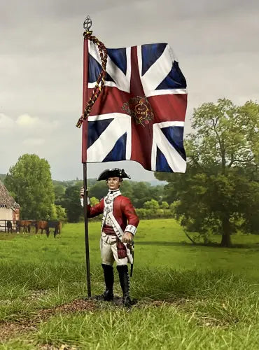 A toy soldier in a red coat uniform stands on grass holding a large British flag with a royal emblem, set against a rural landscape with trees, a fence, a building, and horses in the background.