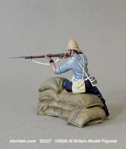 A detailed scale model figurine of a World War I soldier in a crouched position behind sandbags, holding a rifle with a bayonet. The figure is wearing a light blue shirt, dark trousers, and a tan helmet, with a cream shoulder bag and canteen.