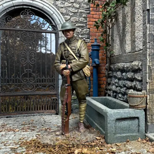 A toy soldier figurine in World War I uniform stands holding a rifle before a stone building and metal gate, with a water trough and blue hand pump nearby.
