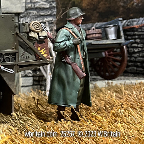 A 1/30 scale toy soldier figurine depicting a U.S. Infantryman from World War II, standing next to a military cart, wearing an Olive Drab raincoat over his uniform and equipment.