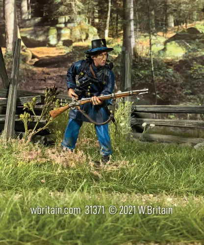 A detailed toy soldier figurine in a blue uniform and hat, holding a rifle, crouches in a grassy area near a wooden fence. The background features an outdoor scene with trees and rocks.