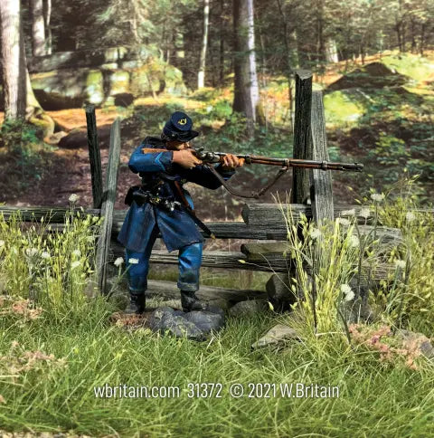 A figurine of a soldier in a blue uniform aims a rifle from behind a wooden fence. The background features a forest scene, and the ground is covered with grass and small plants.