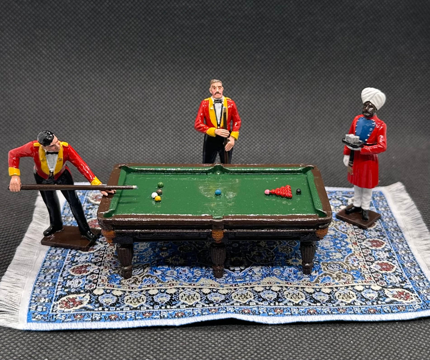 A miniature toy set depicting three figures playing with a small green pool table on a detailed blue and white patterned rug. The figures are dressed in historical attire, with two wearing red jackets and one in a white turban.