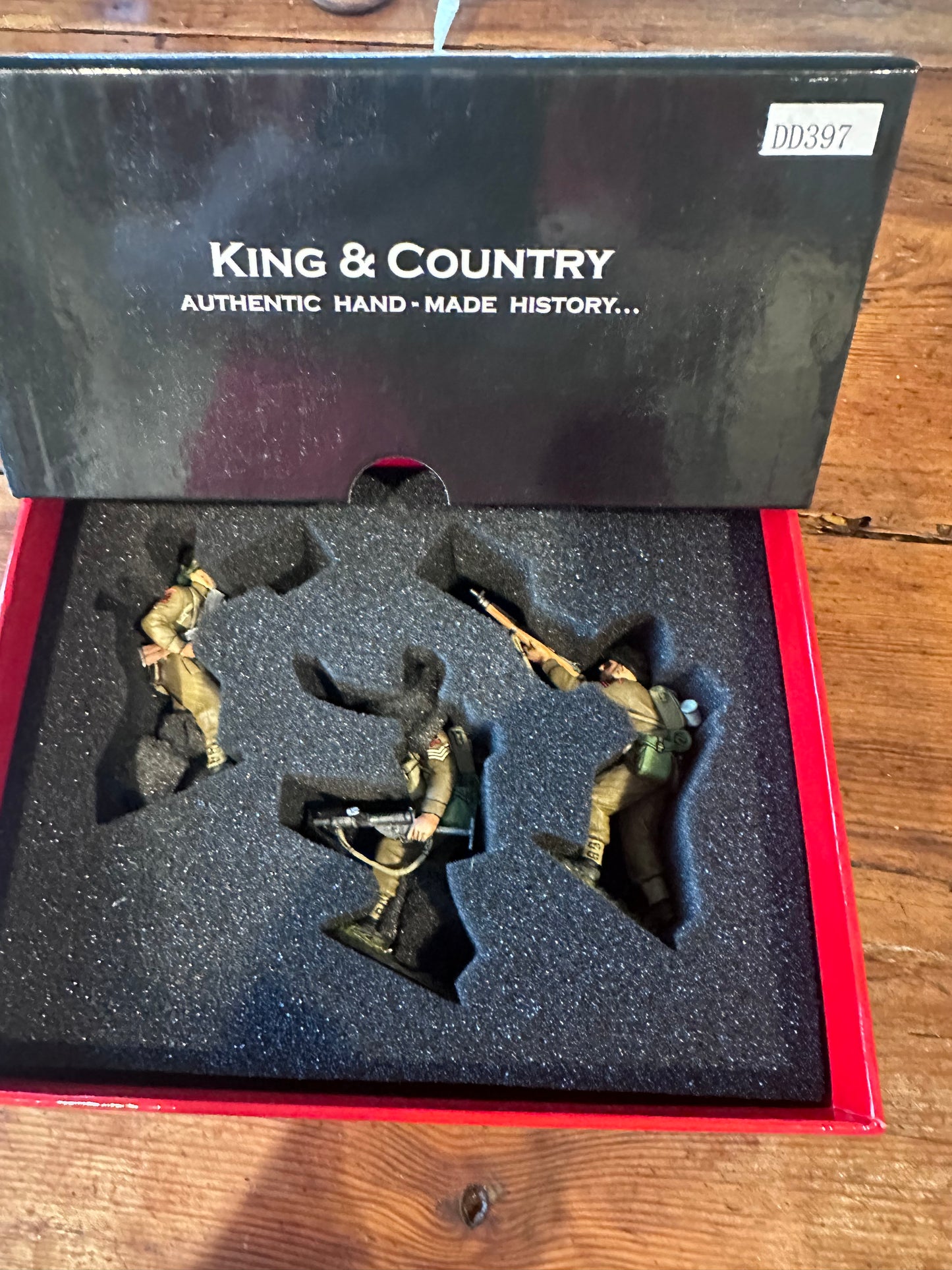 Open black display box for King & Country miniature figures, showing three soldier figurines molded into black foam insert on a red tray, photographed on a wooden surface.