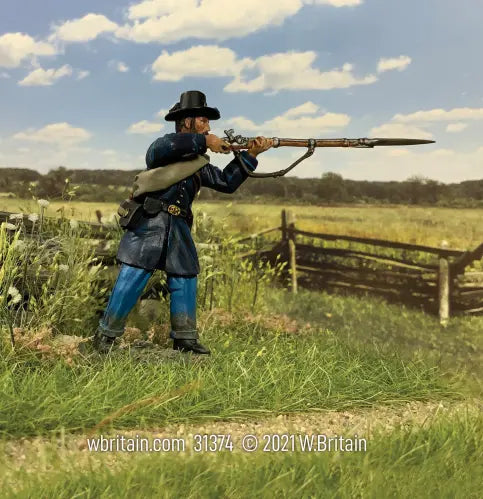 A Federal Iron Brigade Corporal, clad in blue, stands and fires precisely amidst a grassy field. Behind him, a wooden fence and tall grass sway under the sky. At the bottom, W.Britain reminds you of the craftsmanship involved in creating this toy soldier.