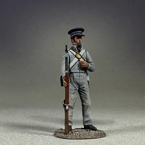 Model figurine of a historical soldier in a blue and gray uniform, holding a rifle with a bayonet. The soldier stands on a textured base, wearing a cap and belt with straps, over a neutral background.