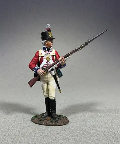 A toy soldier figurine in a red and white uniform stands holding a rifle with a bayonet attached. The soldier wears a tall black hat with a plume, showcasing detailed clothing that suggests a historical military style. The background is plain gray.