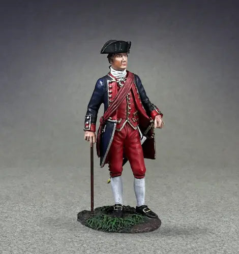 A toy soldier figurine of a colonial-era character wearing a blue coat with red accents, red pants, and a tricorne hat. It stands on a grassy base, holding a book in one hand and a walking stick in the other, gazing thoughtfully to the side.