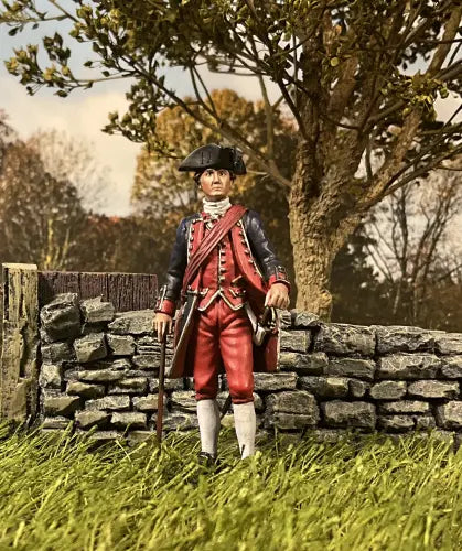 A toy soldier figurine of a colonial-era character wearing a blue coat with red accents, red pants, and a tricorne hat. It stands on a grassy base, holding a book in one hand and a walking stick in the other, gazing thoughtfully to the side. Near a stone wall.