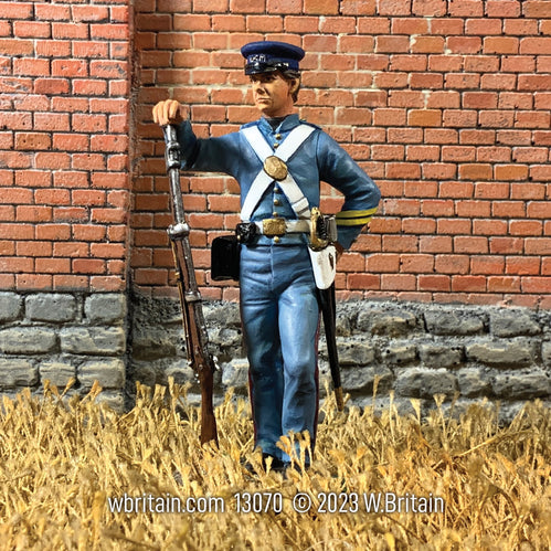 Collectible toy soldier U.S. Marine in fatigue Uniform. He is holding a musket. He is standing outside in front a red brick fence.
