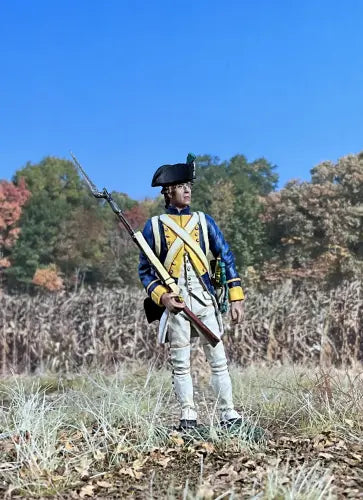 A figurine dressed like a toy soldier in an 18th-century military uniform stands in a grassy field, holding a long rifle with bayonet. He wears a tricorn hat and blue coat with yellow trim, framed by autumn trees under a clear blue sky.