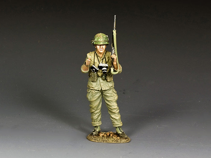 Model of a soldier in green uniform holding rifles and a camera on a gray background