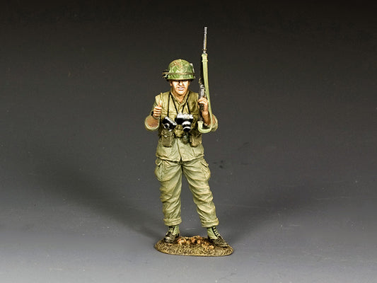 Model of a soldier in green uniform holding rifles and a camera on a gray background