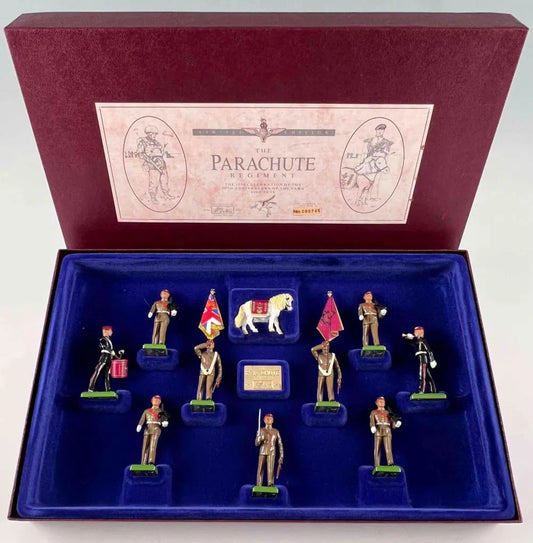 A display box containing ten miniature toy soldiers and a white animal figure, representing The Parachute Regiment. Each soldier holds different flags or items, arranged neatly in a blue velvet-lined box with a description label on the lid.