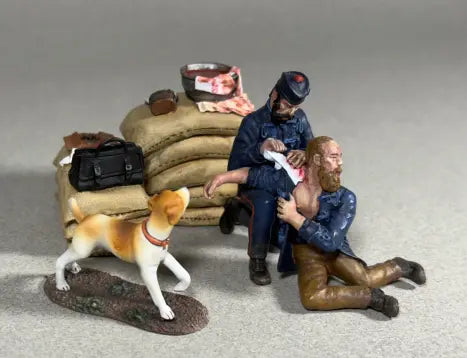 A miniature scene shows a toy soldier medic bandaging a wounded mans arm next to stacked sandbags. A dog stands nearby, and a medical bag with supplies is visible in the background.