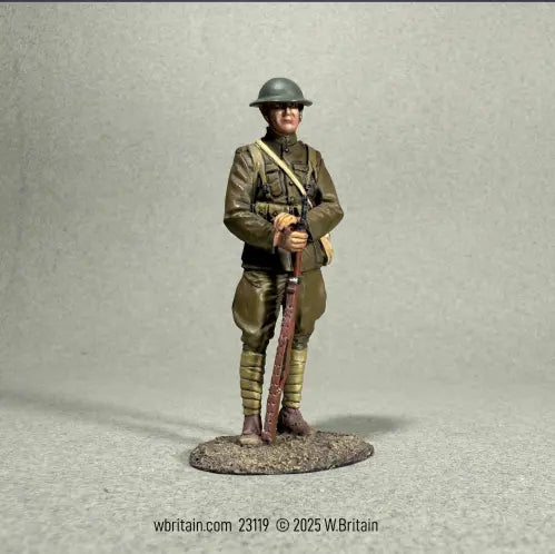 A painted toy soldier figure of a World War I soldier in uniform stands on a textured base, holding a rifle vertically in front of him. The background is plain gray. 