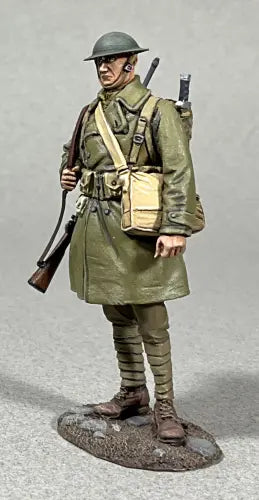 A Toy Soldier figurine of a World War I soldier in a green uniform, helmet, and long coat, carrying a rifle over his shoulder with a beige satchel and gear on his back, standing on a textured base.