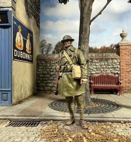 A toy soldier in World War I uniform stands on a cobblestone sidewalk beside a tree, with a brick wall, red bench, and vintage Dubonnet advertisement in the background. The sky is partly cloudy.
