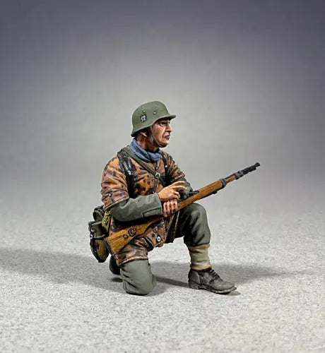 A detailed model figure depicts a soldier kneeling on one knee, wearing a green helmet and camouflage jacket, holding a rifle. The figure is set against a neutral gray background.