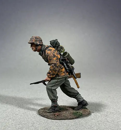 A toy soldier figurine, dressed in a detailed camouflage uniform and helmet, crouches while gripping a rifle. With gear strapped to his back, he is poised on a small textured base against a plain gray backdrop.