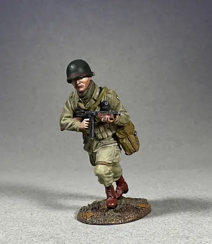 A detailed military miniature of a soldier wearing a green helmet and khaki uniform, holding a rifle and running. This toy soldier has a brown bag slung over one shoulder, standing on a textured base against a simple gray background.