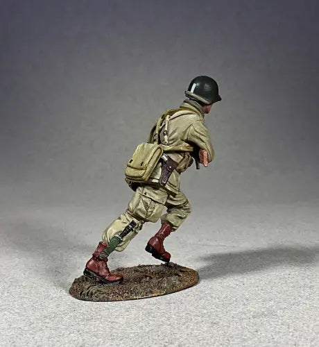 Rear view of A detailed military miniature of a soldier wearing a green helmet and khaki uniform, holding a rifle and running. This toy soldier has a brown bag slung over one shoulder, standing on a textured base against a simple gray background.