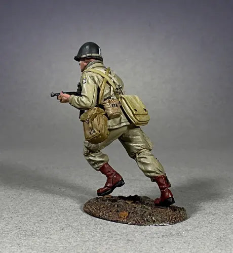 Side view of  A detailed military miniature of a soldier wearing a green helmet and khaki uniform, holding a rifle and running. This toy soldier has a brown bag slung over one shoulder, standing on a textured base against a simple gray background.