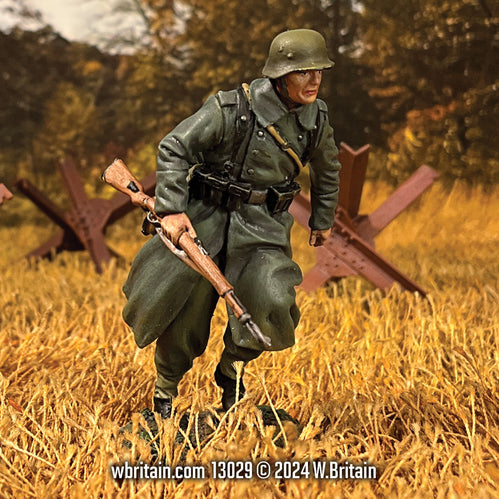 Miniature figurine of a German Grenadier running through a field, clutching a Karabiner 98K rifle. Dressed in a long green greatcoat and helmet, his expression focused, he moves swiftly across a golden autumn landscape with anti-tank obstacles in the background, capturing the urgency of battlefield maneuvers during World War II.