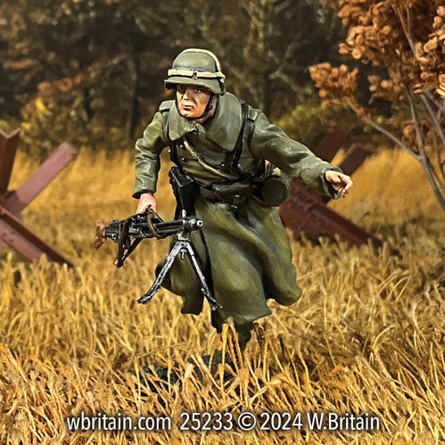 Miniature figurine of a German Grenadier running through an autumn field, holding an MG 42 machine gun. He is dressed in a long green greatcoat and helmet, his expression focused as he advances through the golden, grassy terrain. The detailed figurine captures the urgency and mobility required in the battlefield conditions of World War II.