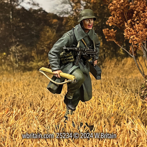 Miniature figurine of a German Grenadier running through a golden autumn field, dressed in a greatcoat and helmet, carrying a spare MG 42 machine gun barrel and a metal case. His expression is intense, highlighting the urgency of battlefield operations during World War II.