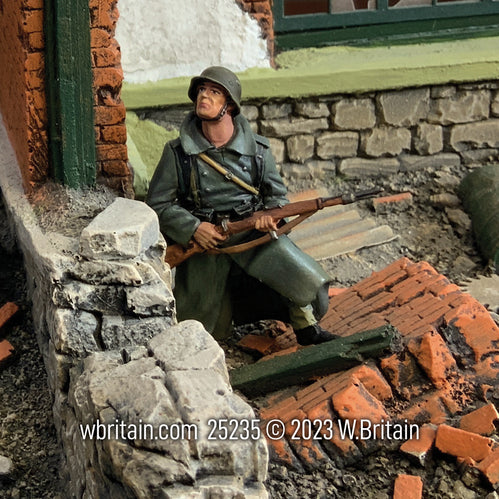 Miniature figurine of a German Grenadier in a greatcoat, kneeling and looking up, positioned against a war-damaged urban backdrop. The soldier, in a dark green uniform with a helmet, holds a rifle and is surrounded by broken bricks and rubble, indicating recent combat.