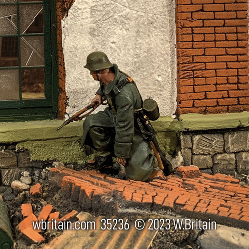 A detailed miniature figurine of a German Grenadier kneeling in a greatcoat, set against a wartime urban backdrop. The soldier is depicted with a focused expression, holding a rifle and dressed in a dark green military uniform with a helmet.