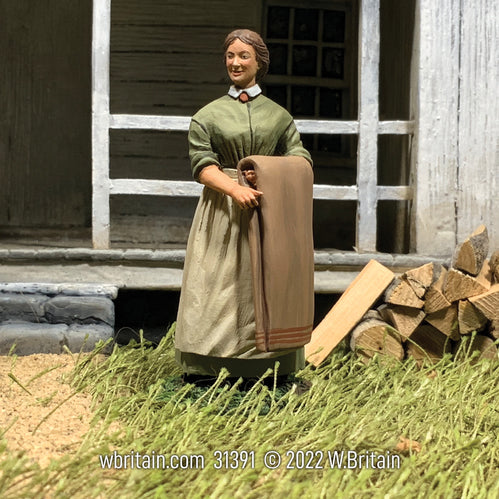 Civilian miniature Figure of a lady with long dress and folded blanket. She is standing in front of an old home.