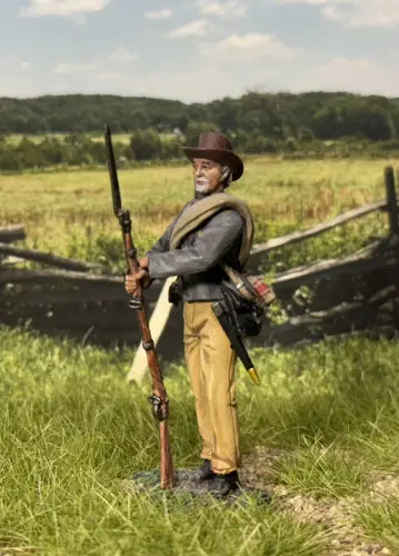 Confederate Infantry at Rest. Figure of a Civil War soldier holding a rifle in a grassy field with a wooden fence and trees in the background.