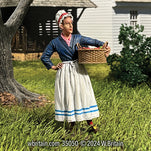 A highly detailed miniature figurine of a late 18th-century woman named Mary, holding a large wicker basket filled with fresh strawberries. She is dressed in a traditional outfit with a blue jacket, white skirt accented with blue stripes at the hem, and a red and white bonnet. Mary stands in a grassy field beside a wooden house, her gaze directed off into the distance, capturing a serene moment of rural life