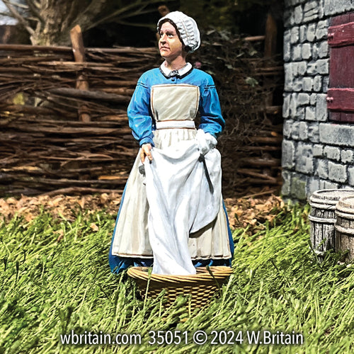 A highly detailed miniature figurine of a woman named Vivian, depicted in the mid-19th century, holding clean linens. She wears a traditional blue dress with a white apron and bonnet, standing next to a wicker basket in a grassy field. Behind her is a rustic wooden fence and a stone building, reflecting the rural setting typical of a bleachfield used for sun-whitening textiles. Vivian looks contemplatively to the side, capturing a moment of daily life during the Industrial Revolution.