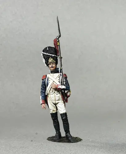 A toy soldier figurine of a soldier in historical military uniform stands against a gray background. The uniform includes a black hat with a red plume, a blue coat with white lapels, and white trousers. The soldier holds a long rifle with a bayonet, showcasing the detailed craftsmanship.