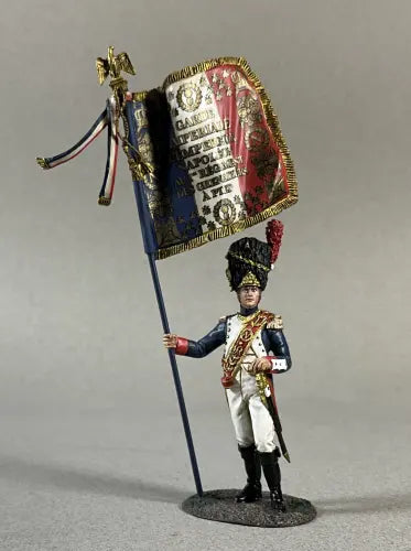 A toy soldier figurine stands proudly in a historic military uniform, gripping a tall flagpole. The vibrant flag displays text and ornamental designs. The uniform features a dark blue coat, white trousers, and a decorated hat adorned with striking red and black feathers.