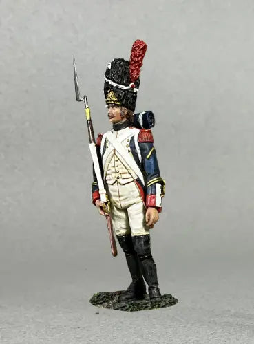 A miniature figurine of a Napoleonic soldier wearing a tall black bearskin hat with a red plume, a blue uniform with white trousers, and black boots, holding a rifle with a bayonet, standing against a gray background.