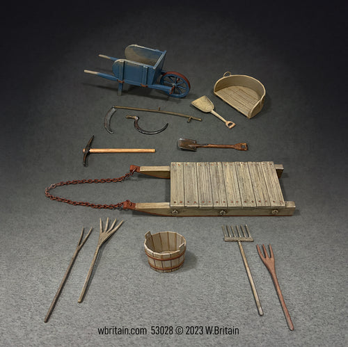 The set includes a blue wheelbarrow, two pitchforks, a scythe, a sickle, a stone boat sled, a winnowing tray, a wooden grain shovel, and a few other traditional farming implements.