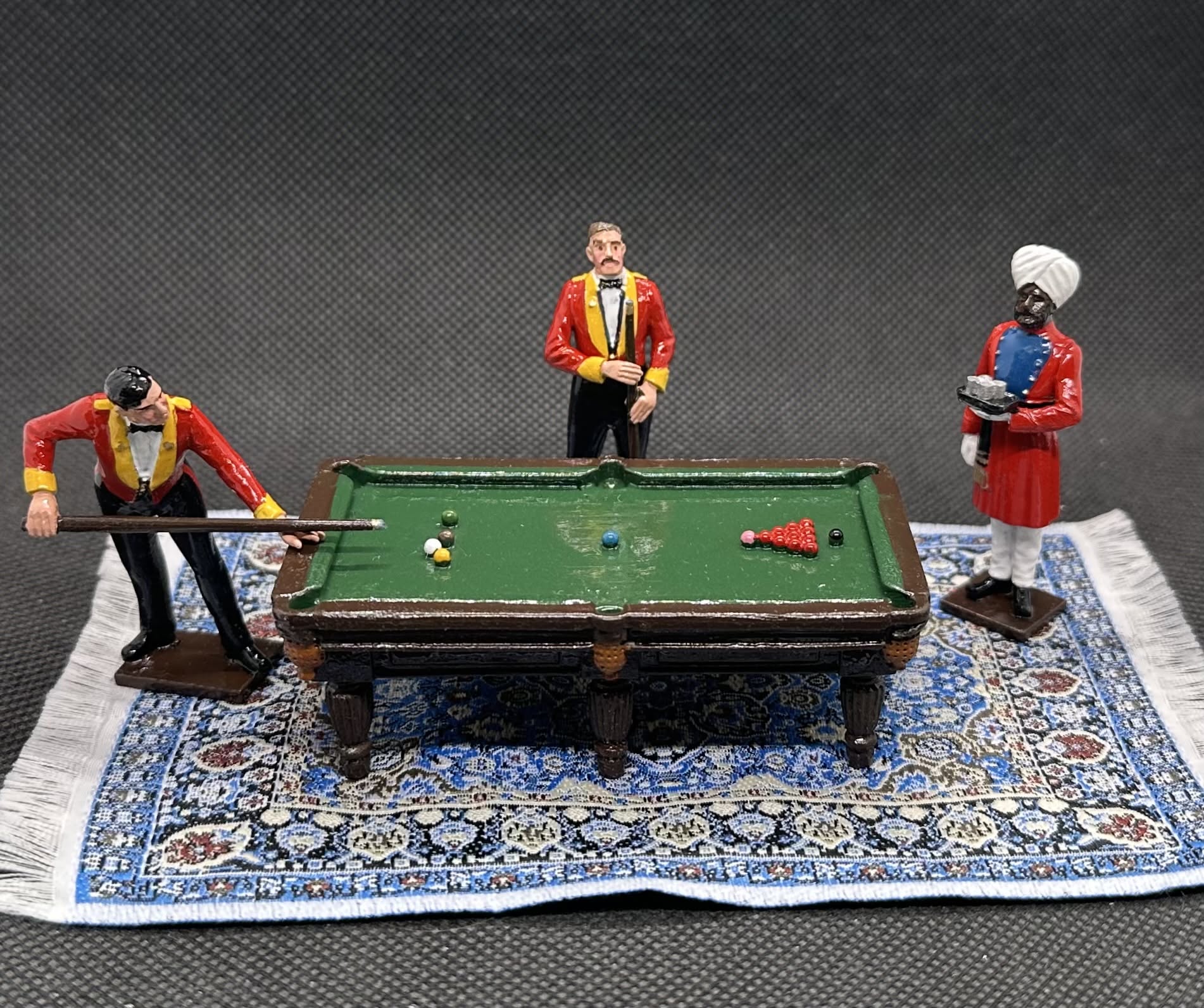 A miniature toy set depicting three figures playing with a small green pool table on a detailed blue and white patterned rug. The figures are dressed in historical attire, with two wearing red jackets and one in a white turban.