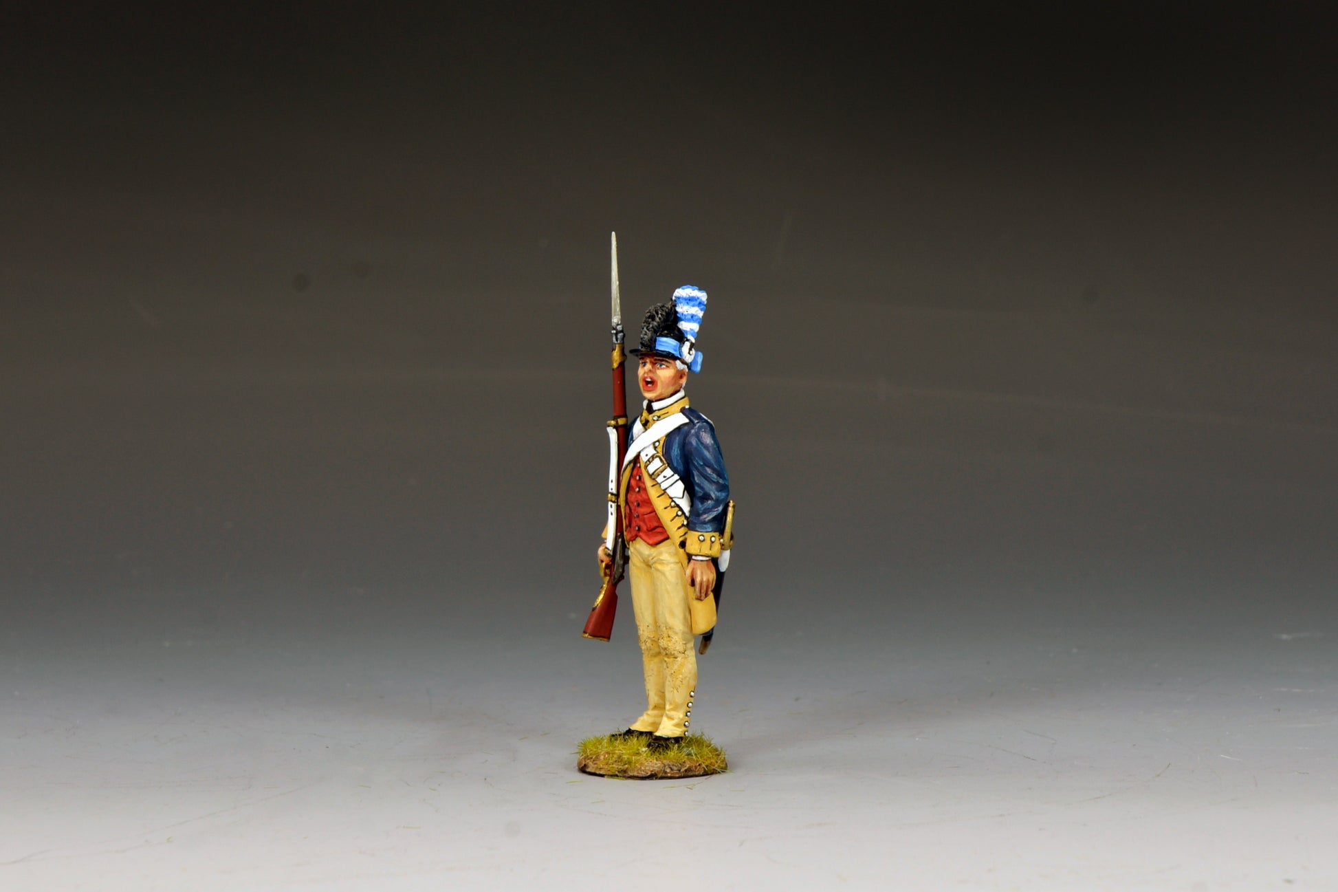 A figurine of a Revolutionary War soldier stands at attention, wearing a blue and red uniform with tan trousers, white crossbelts, and a tall black hat with a blue plume, holding a musket upright by his side.