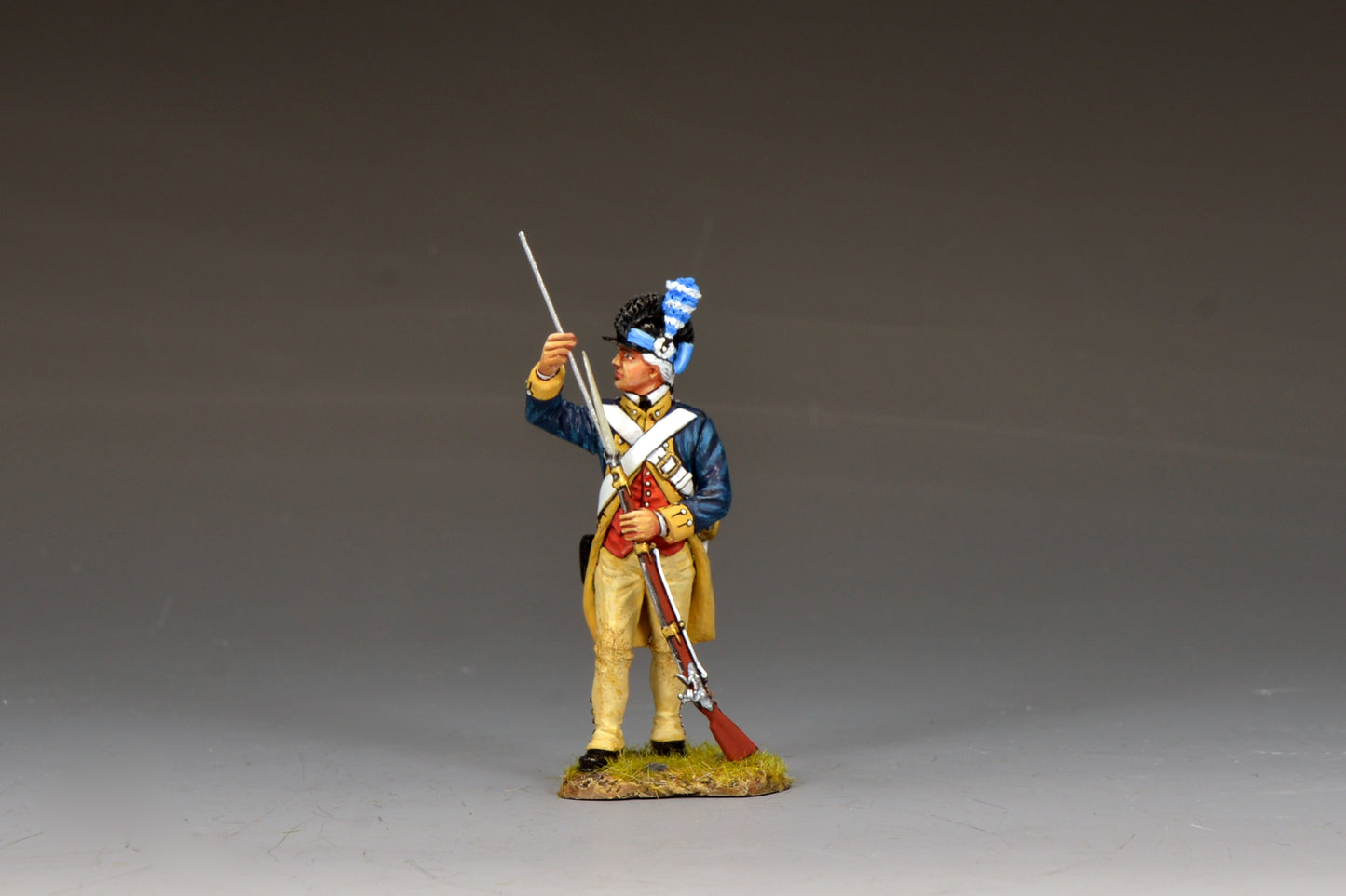 A detailed figurine of an 18th-century soldier in a blue and yellow uniform, holding a rifle upright with one hand and saluting with the other, stands on a small grassy base against a plain background.