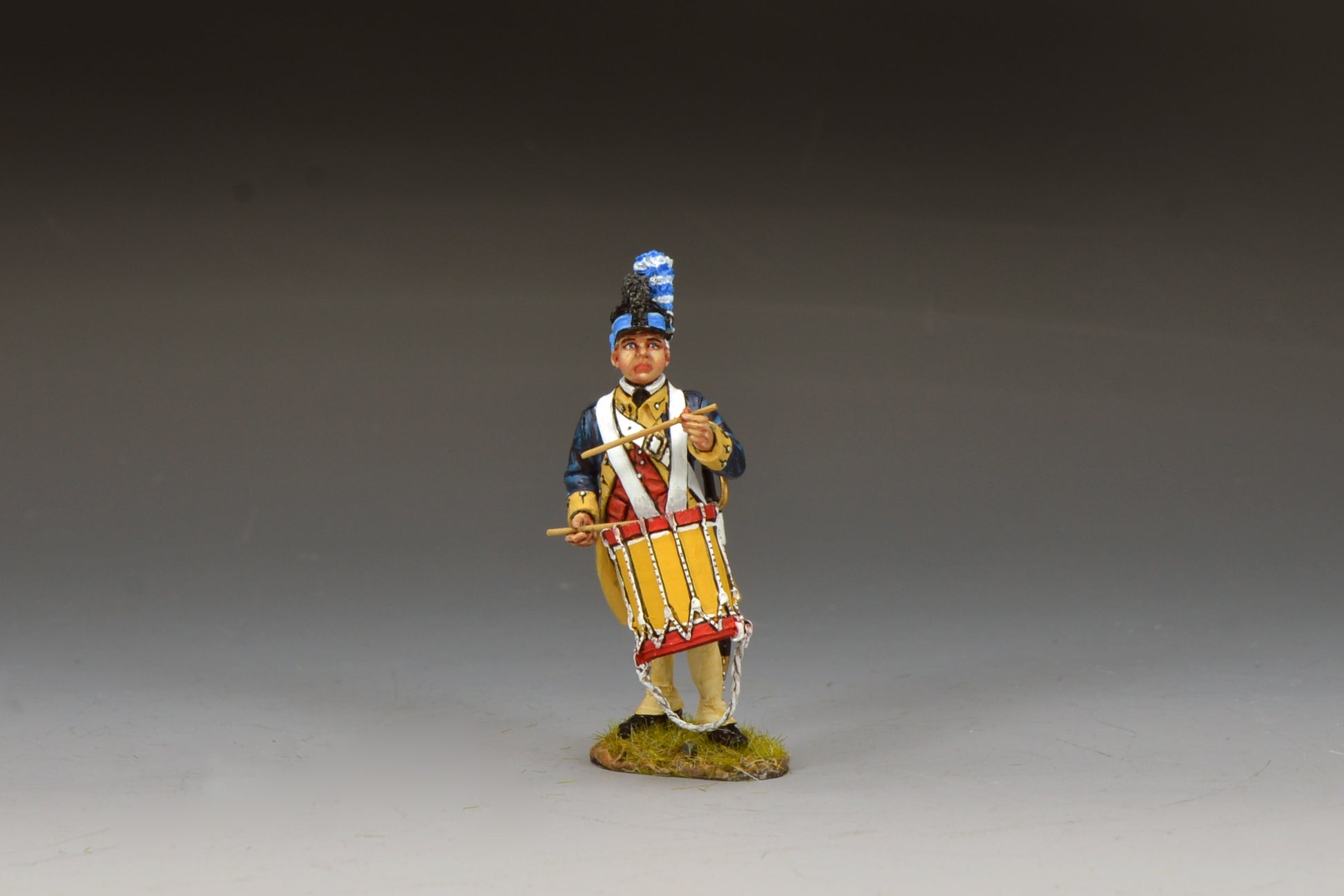 A painted miniature figure of a historical soldier in uniform, playing a drum. The figure stands on a small grassy base and wears a dark blue coat, cream pants, and a tall hat with a blue plume.