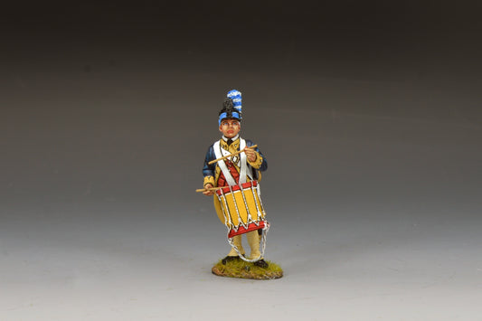A painted miniature figure of a historical soldier in uniform, playing a drum. The figure stands on a small grassy base and wears a dark blue coat, cream pants, and a tall hat with a blue plume.