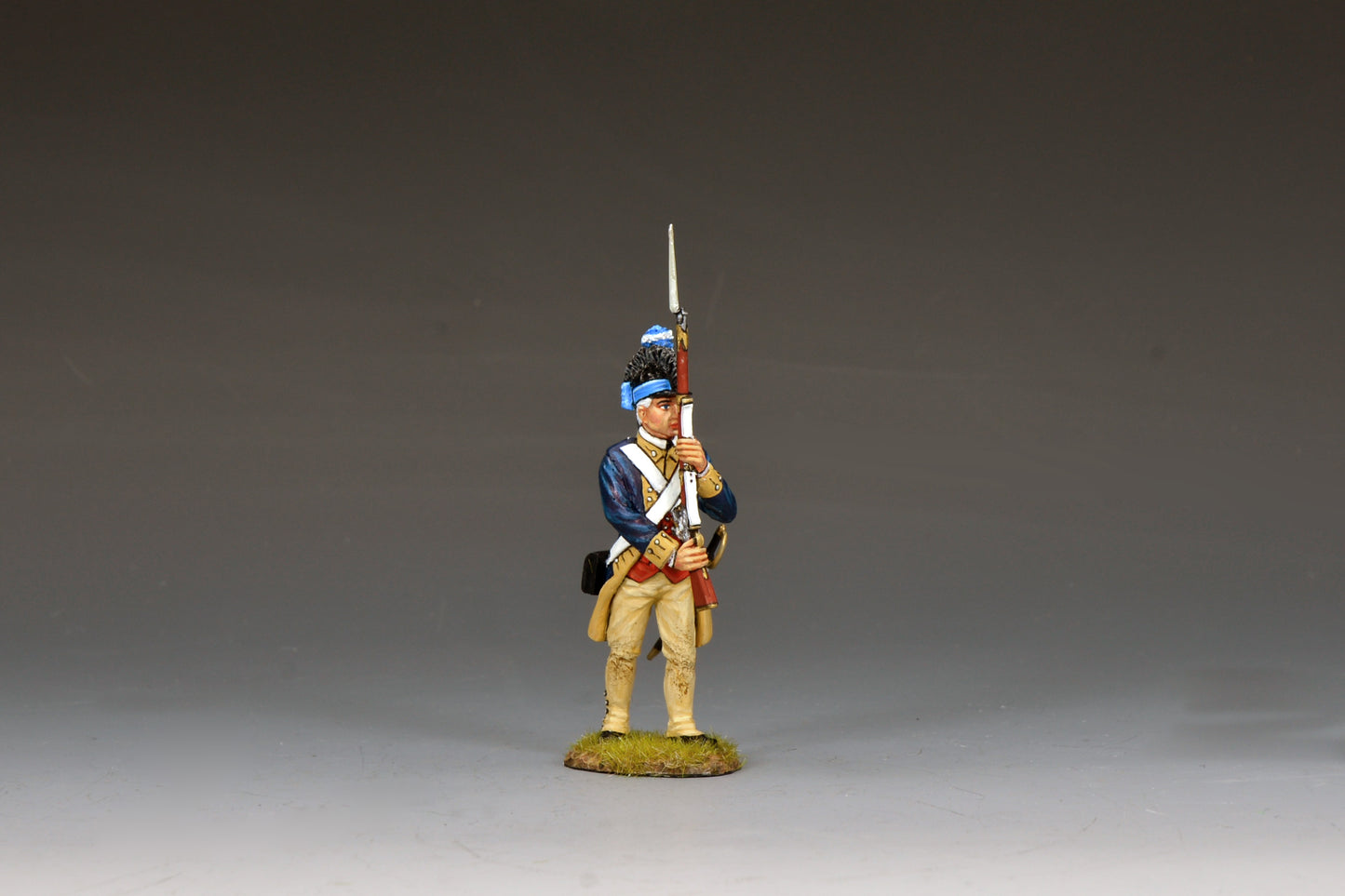 A painted miniature figurine of a Revolutionary War soldier in blue and tan uniform holding a musket upright, standing on a small patch of grass against a plain gray background.