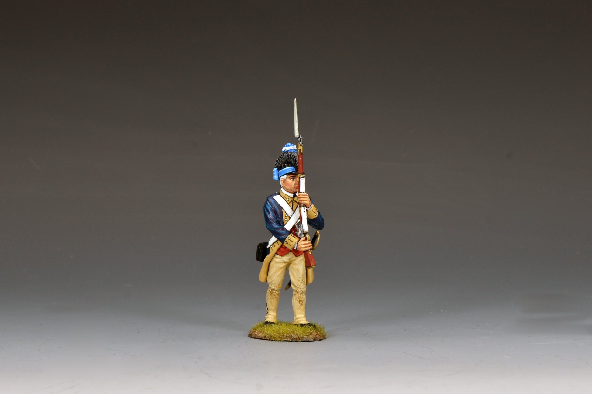 A painted miniature figurine of a Revolutionary War soldier in blue and tan uniform holding a musket upright, standing on a small patch of grass against a plain gray background.