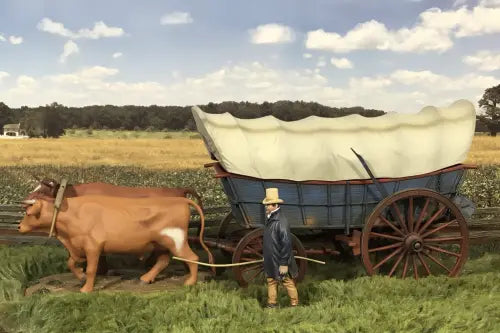 Model of a covered wagon being pulled by a cow with a person standing nearby, set against a scenic background.