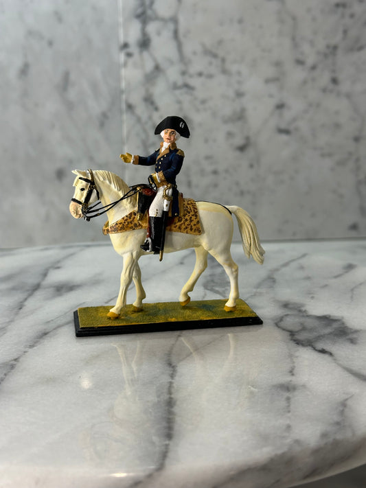 Handcrafted metal figurine of General George Washington on horseback, painted in detail. Washington is depicted in a blue and gold military uniform with a black tricorn hat, riding a white horse adorned with a leopard-print saddle cloth. The figurine is set against a marbled gray background, standing on a yellowish-green base.