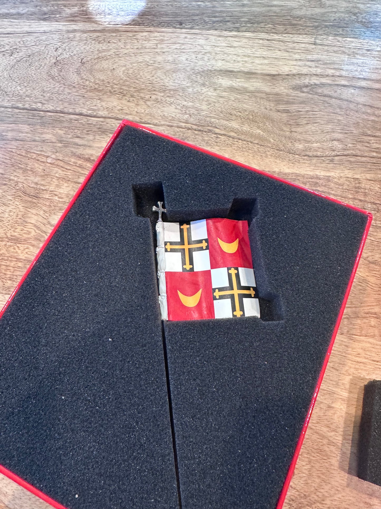 Small flag with crosses and moons on a black display stand with red border, on a wooden surface. The Teutonic Flagbearer